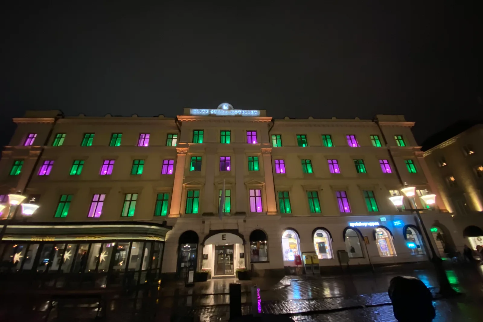 Stor ljus byggnad med fönster som lyser gröna och lila, blöt torgyta framför